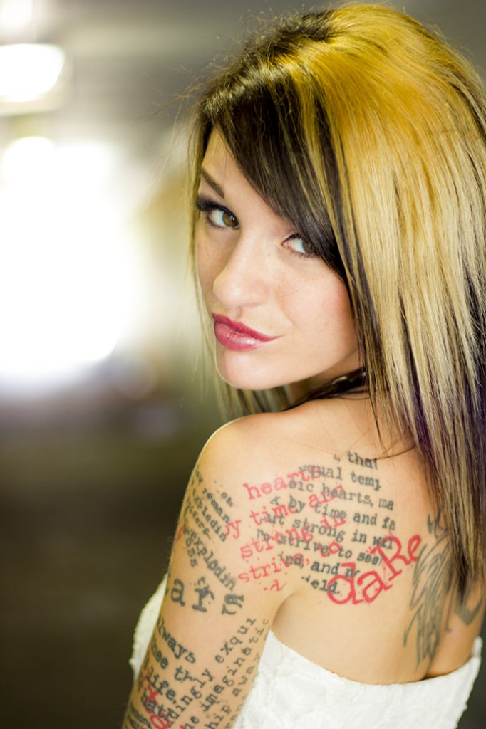 Portrait photo of Emily Wood by Sugar Cube Entertainment. A young woman with typewriter-style text tattoos on her shoulder, back and arm, looking back at the camera over her shoulder.