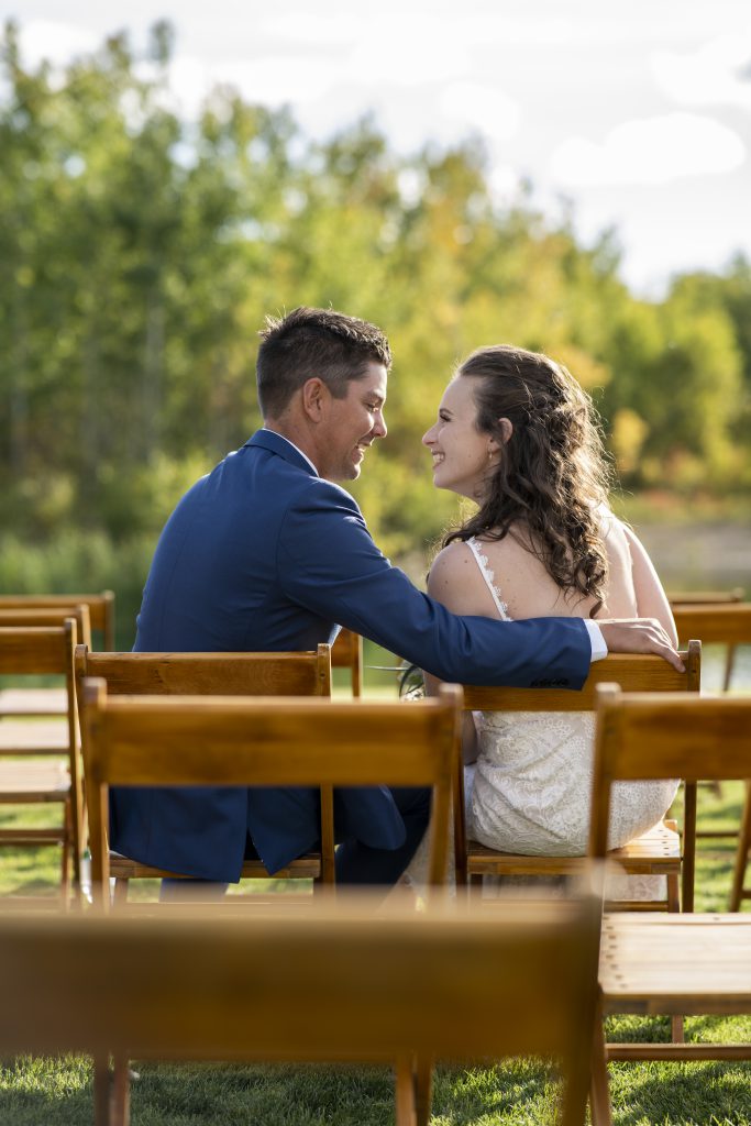 Groom and bride sitting in chairs looking at each other with smiles at their wedding